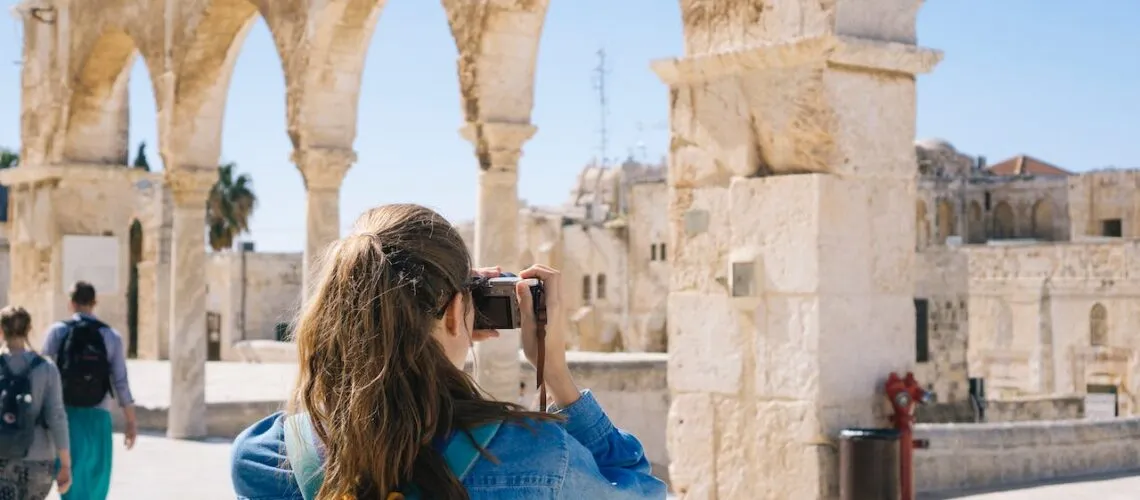 A tourist photographs ancient stone arches under a clear blue sky, capturing historic architecture.