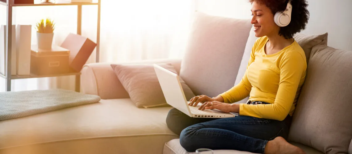 A person wearing headphones works on a laptop while seated on a sofa in a well-lit room.