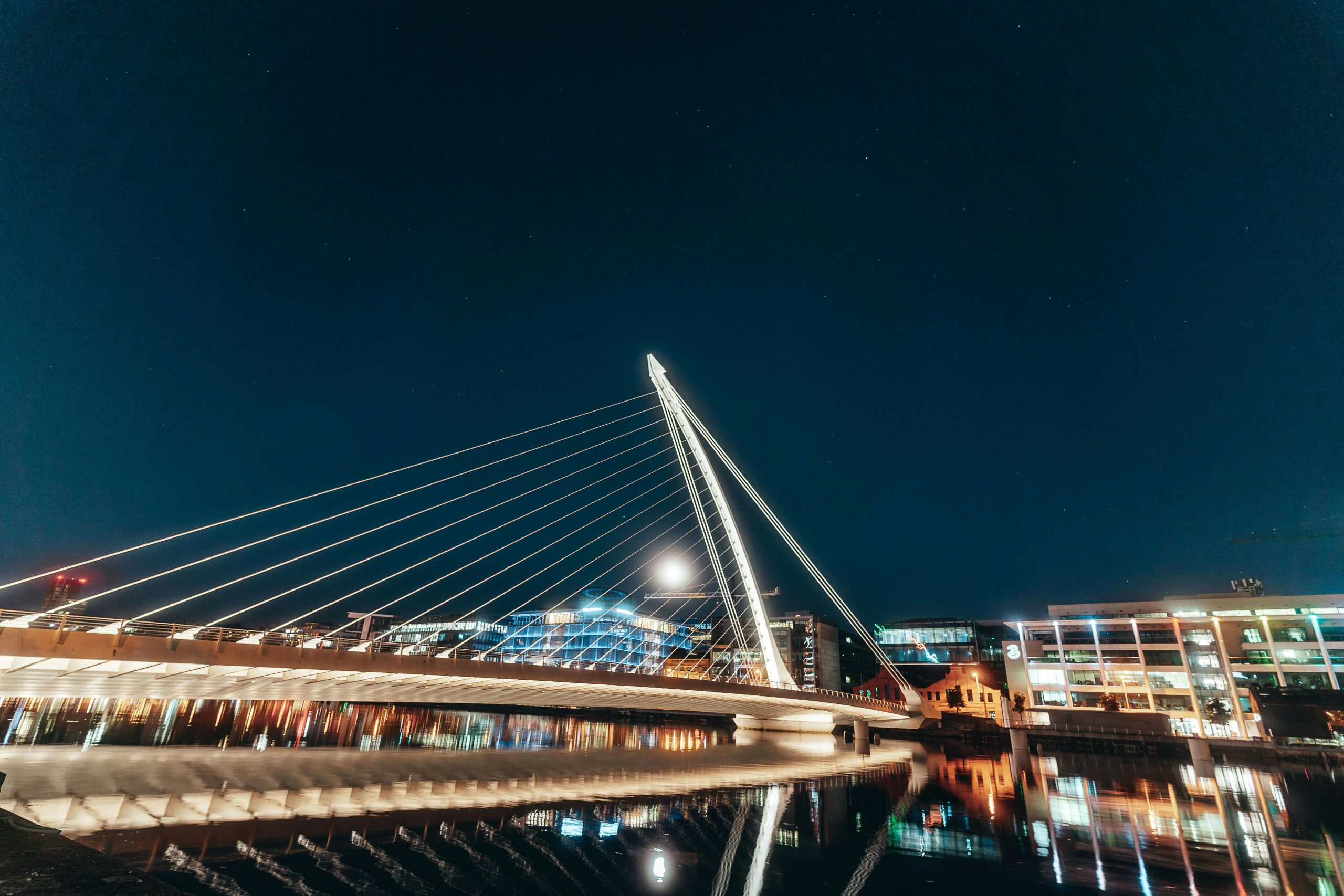 Night view of Samuel Beckett Bridge in Dublin, illuminated over calm waters with city lights.