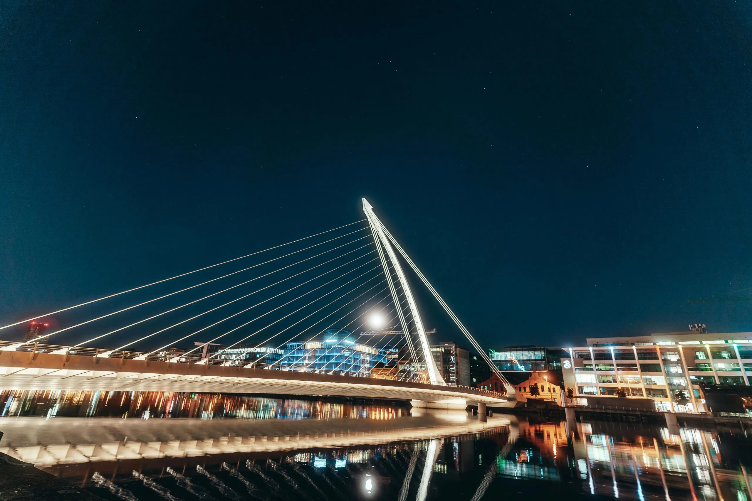 Night view of Samuel Beckett Bridge in Dublin, illuminated over calm waters with city lights.