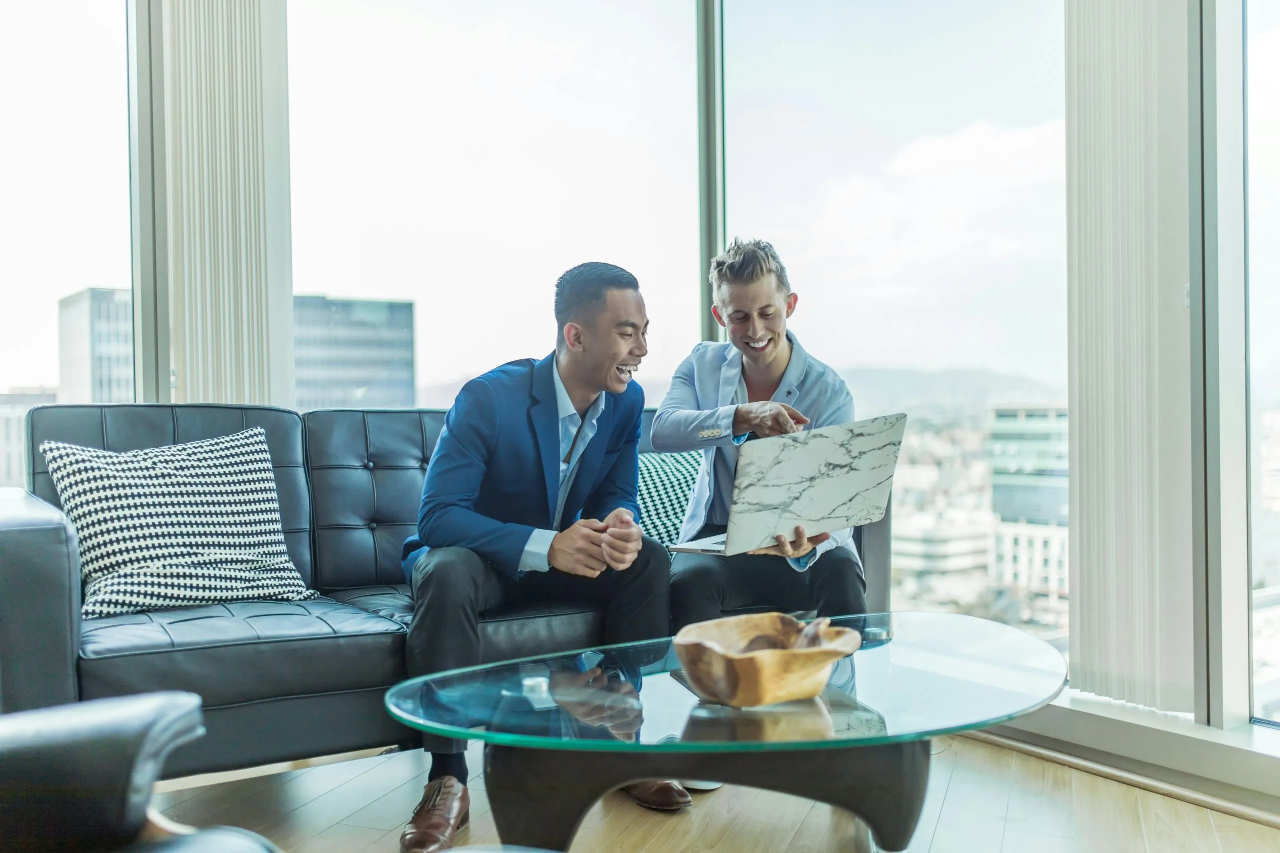 Two professionals collaborate in a modern office, seated on a sofa with a laptop and glass table nearby.