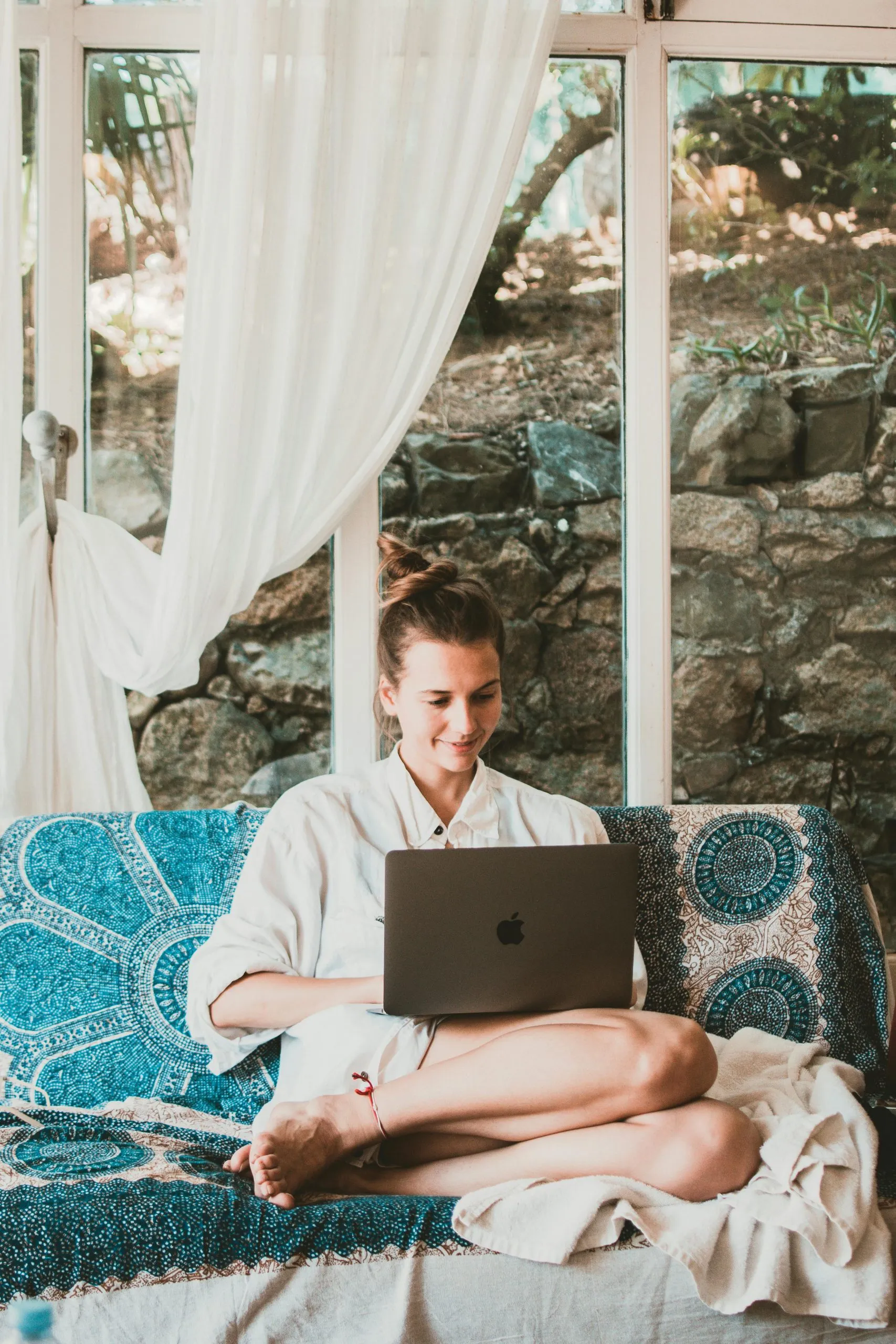 A person sits cross-legged on a patterned couch, working on a laptop in a bright, serene setting.