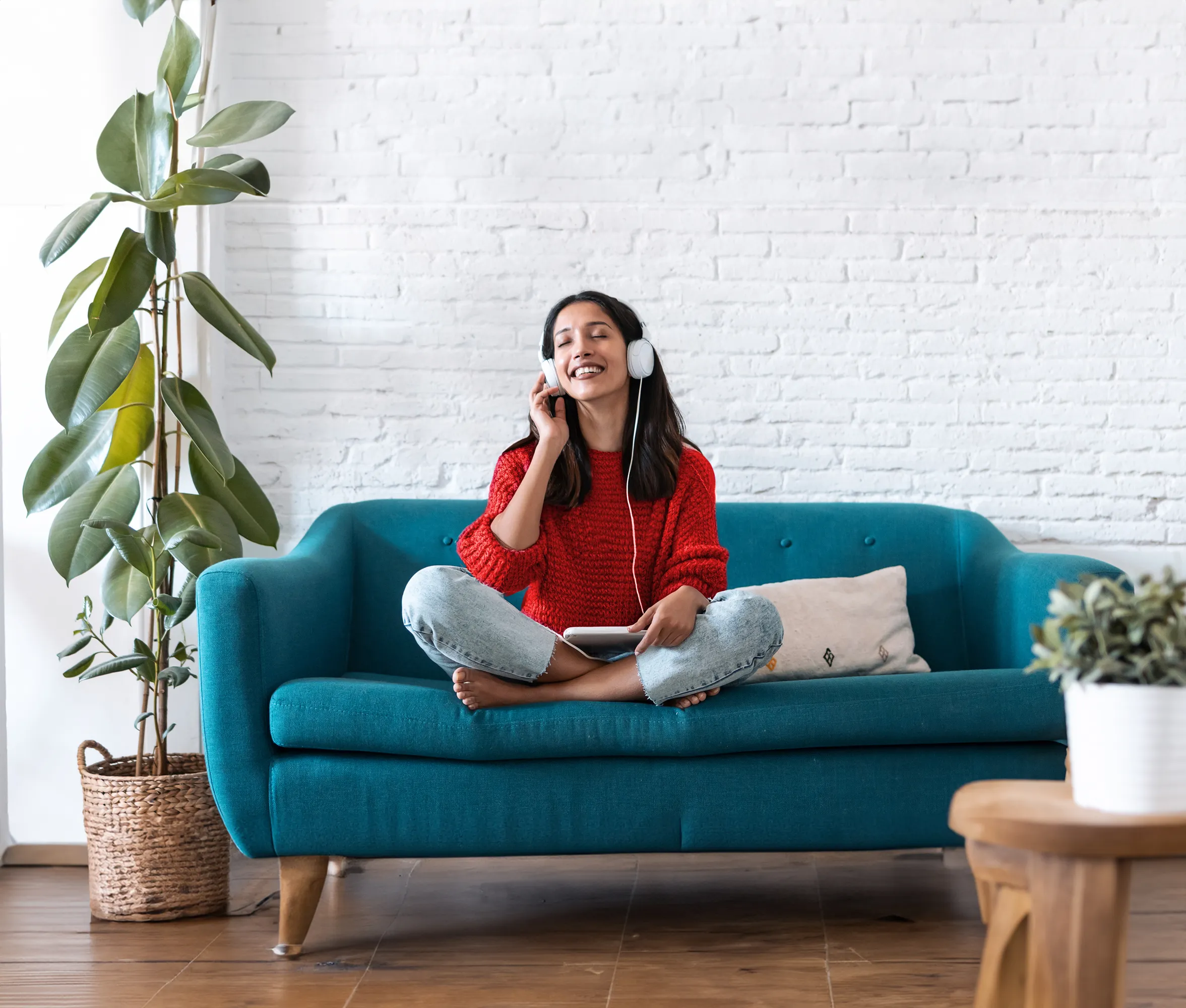 A person in a red sweater sits cross-legged on a blue sofa, wearing headphones and holding a tablet.