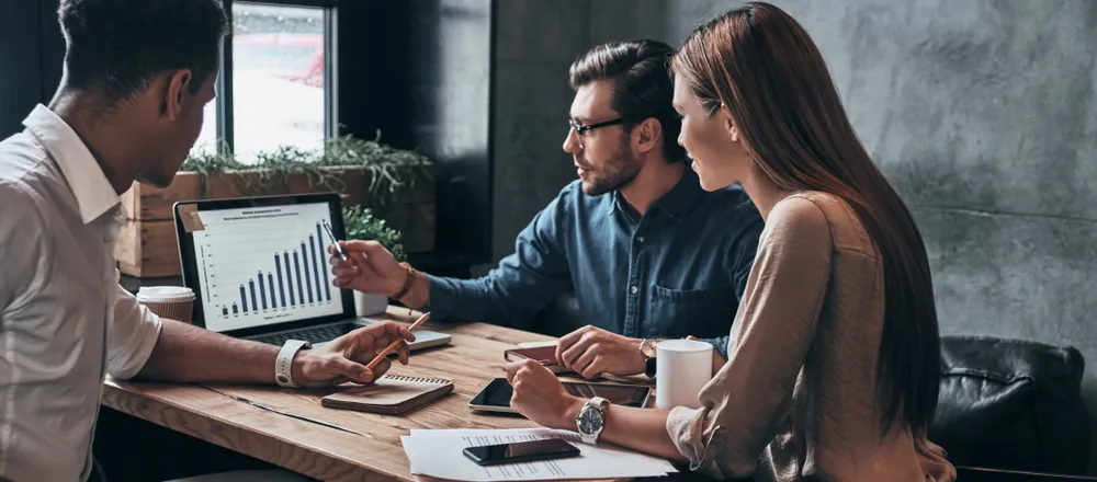 Three colleagues discuss data displayed on a laptop, surrounded by notebooks and coffee cups.