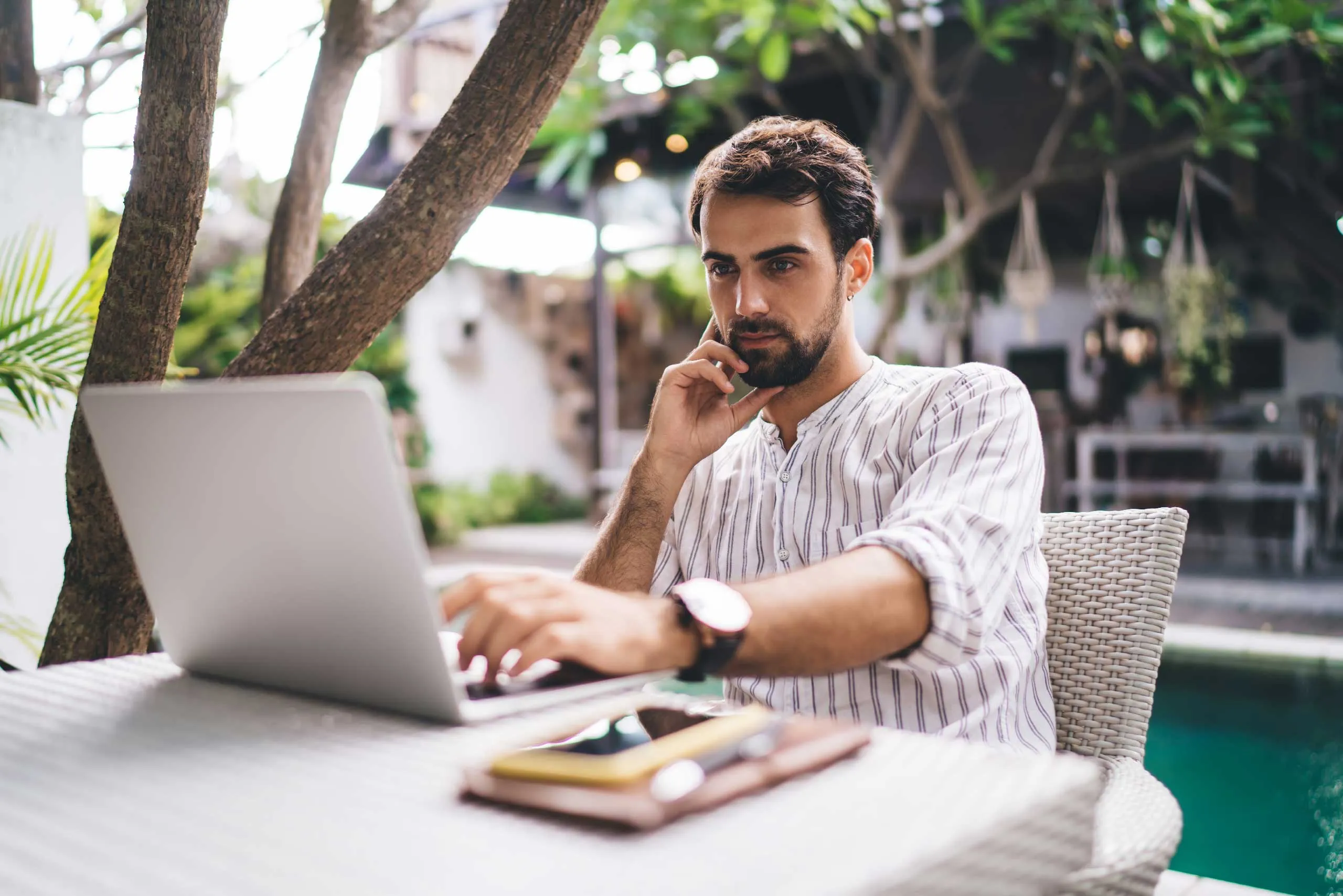 A man works on a laptop outdoors near a pool, surrounded by greenery and a relaxed setting.
