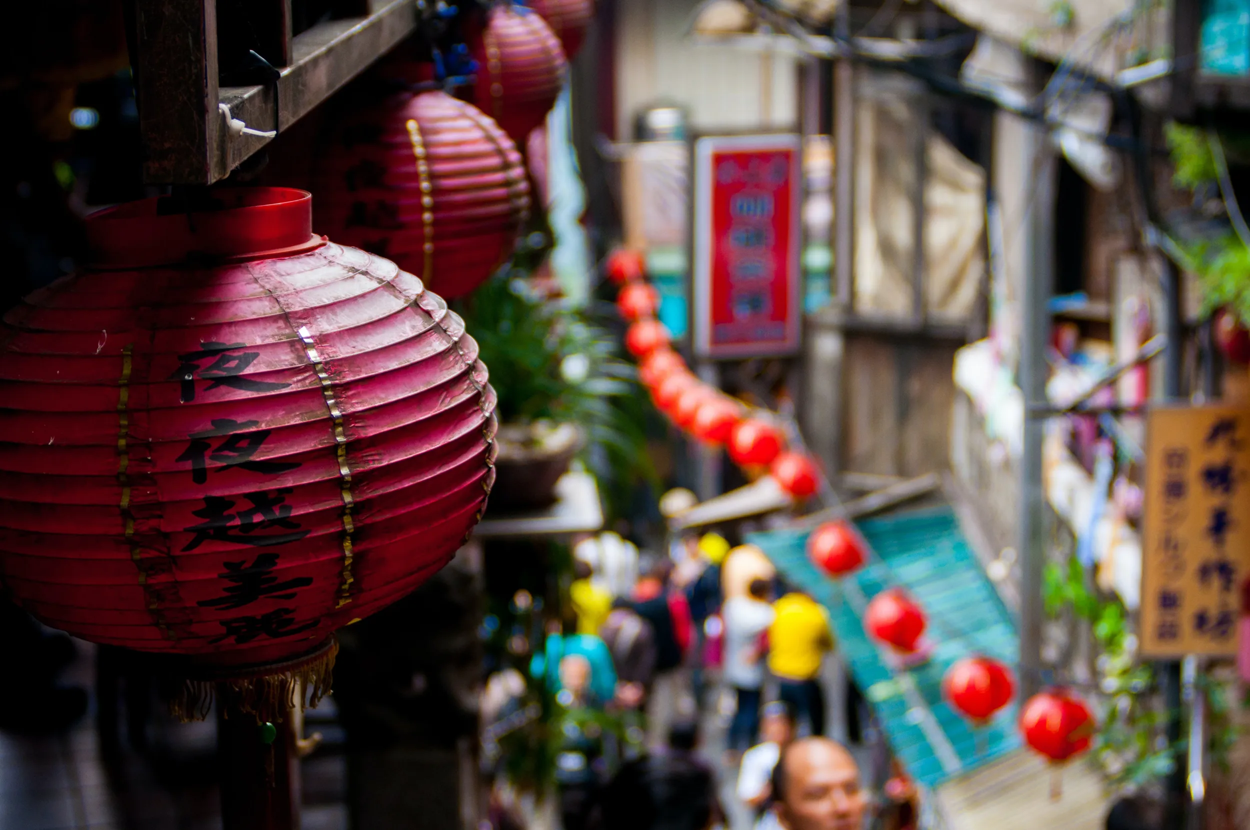 Red lanterns with Chinese characters hang above a bustling, colorful street in an urban setting.