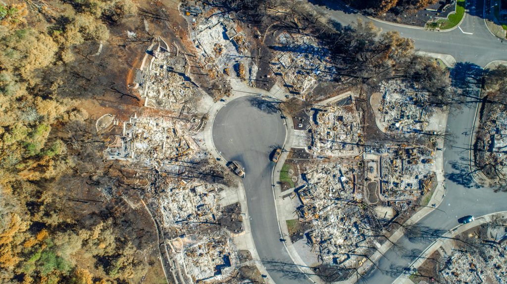 Aerial view of a cul-de-sac with homes destroyed by a wildfire, surrounded by scorched trees.