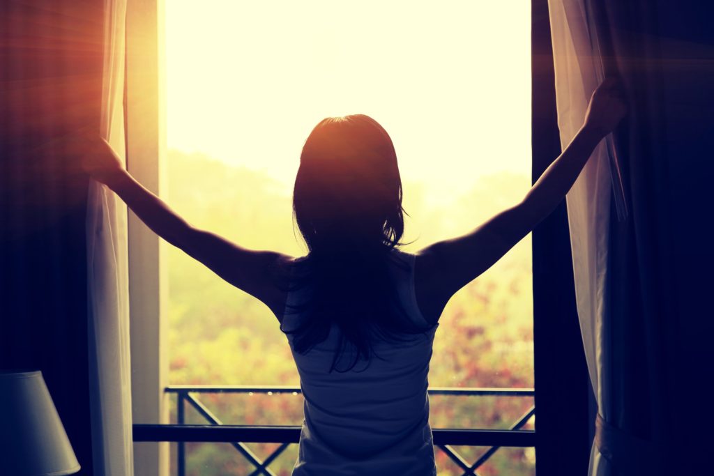 A woman stands by a window, opening curtains to let in sunlight, with a serene outdoor view.