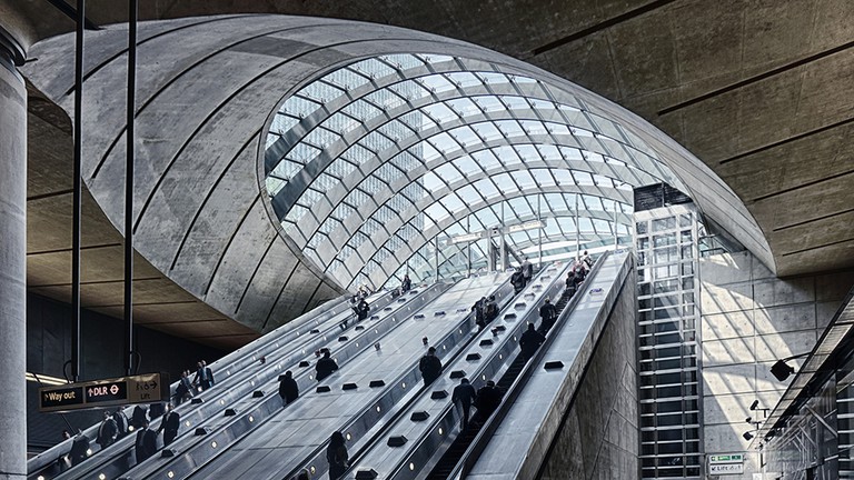 Modern escalators ascend under a curved glass roof in a spacious, concrete urban transit station.