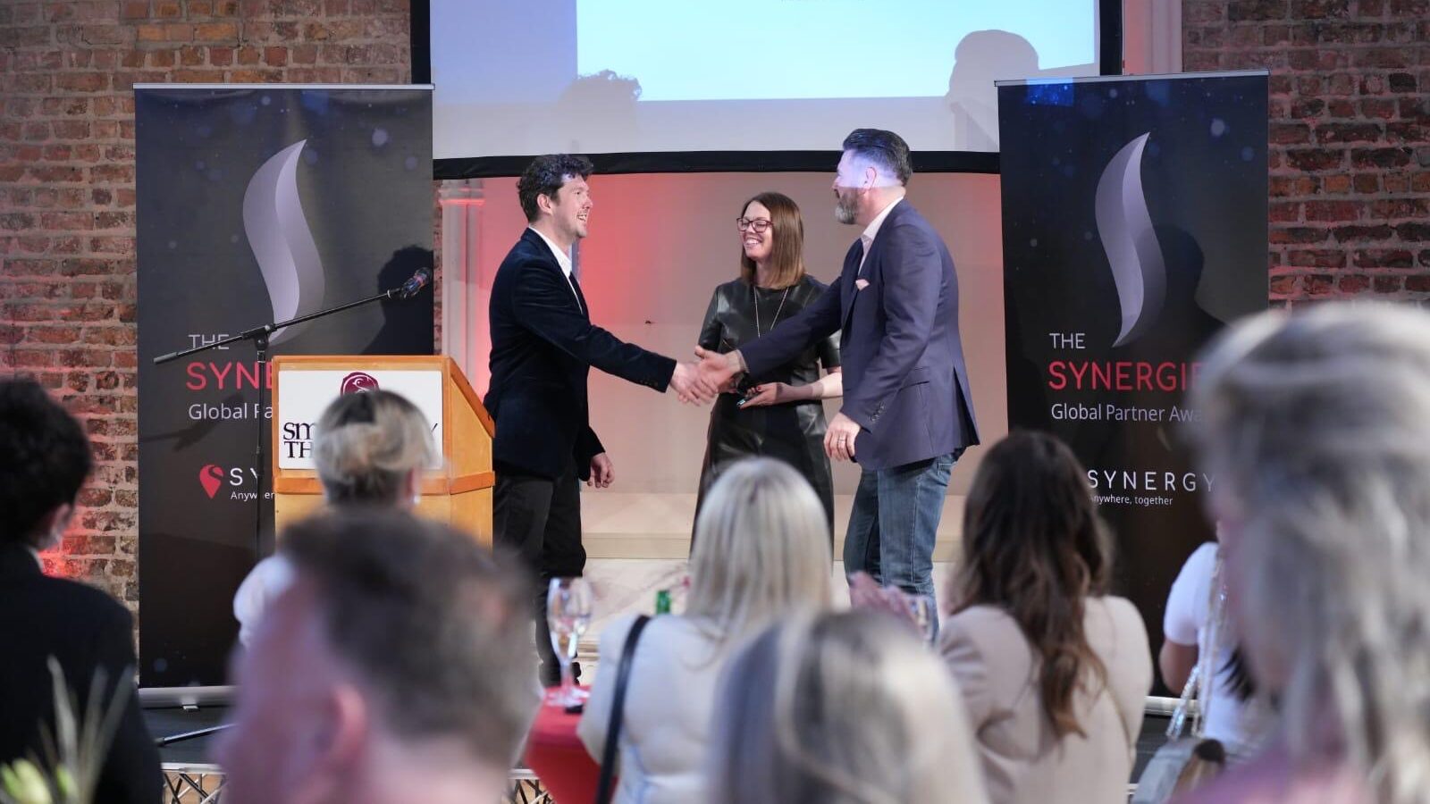 Two individuals shake hands on stage at the Synergize Global Partner Awards event.
