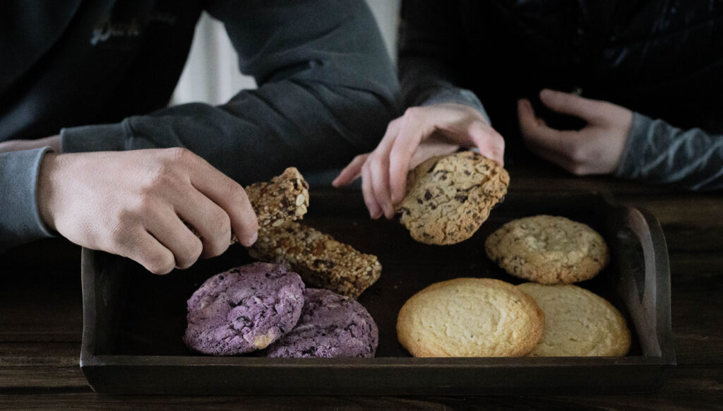 Hands reaching for assorted cookies and bars on a wooden tray, showcasing variety and texture.