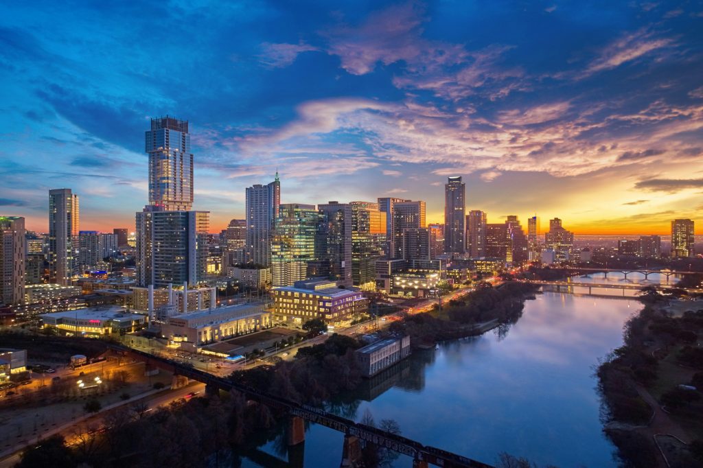 A vibrant city skyline at sunset, reflecting on a calm river with illuminated buildings.