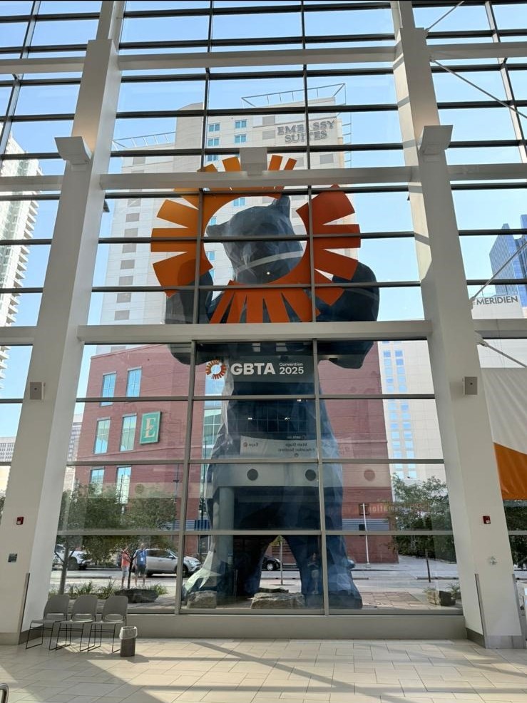 A large blue bear sculpture peers into a glass building, framed by a GBTA 2025 convention logo.