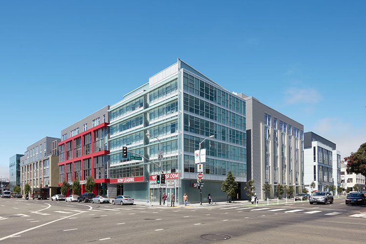 Modern multi-story mixed-use buildings line a busy urban intersection under a clear blue sky.