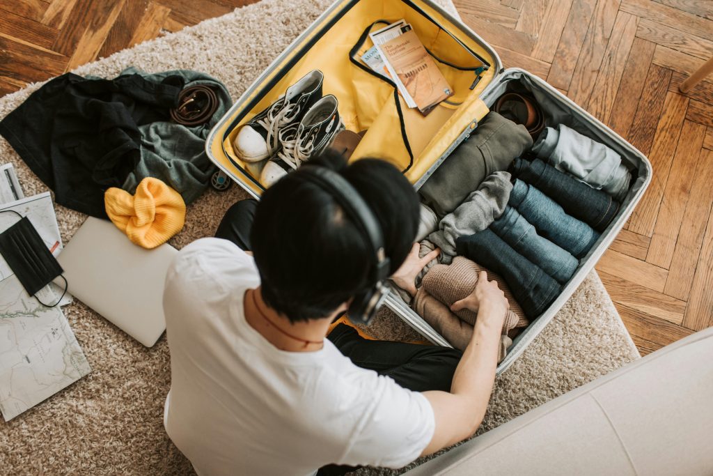 A person packs a suitcase with neatly folded clothes, shoes, and travel essentials on a wooden floor.