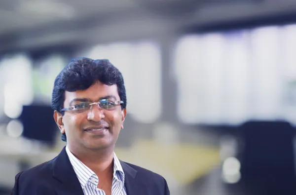 A man in formal attire stands in a modern office setting with a blurred background.