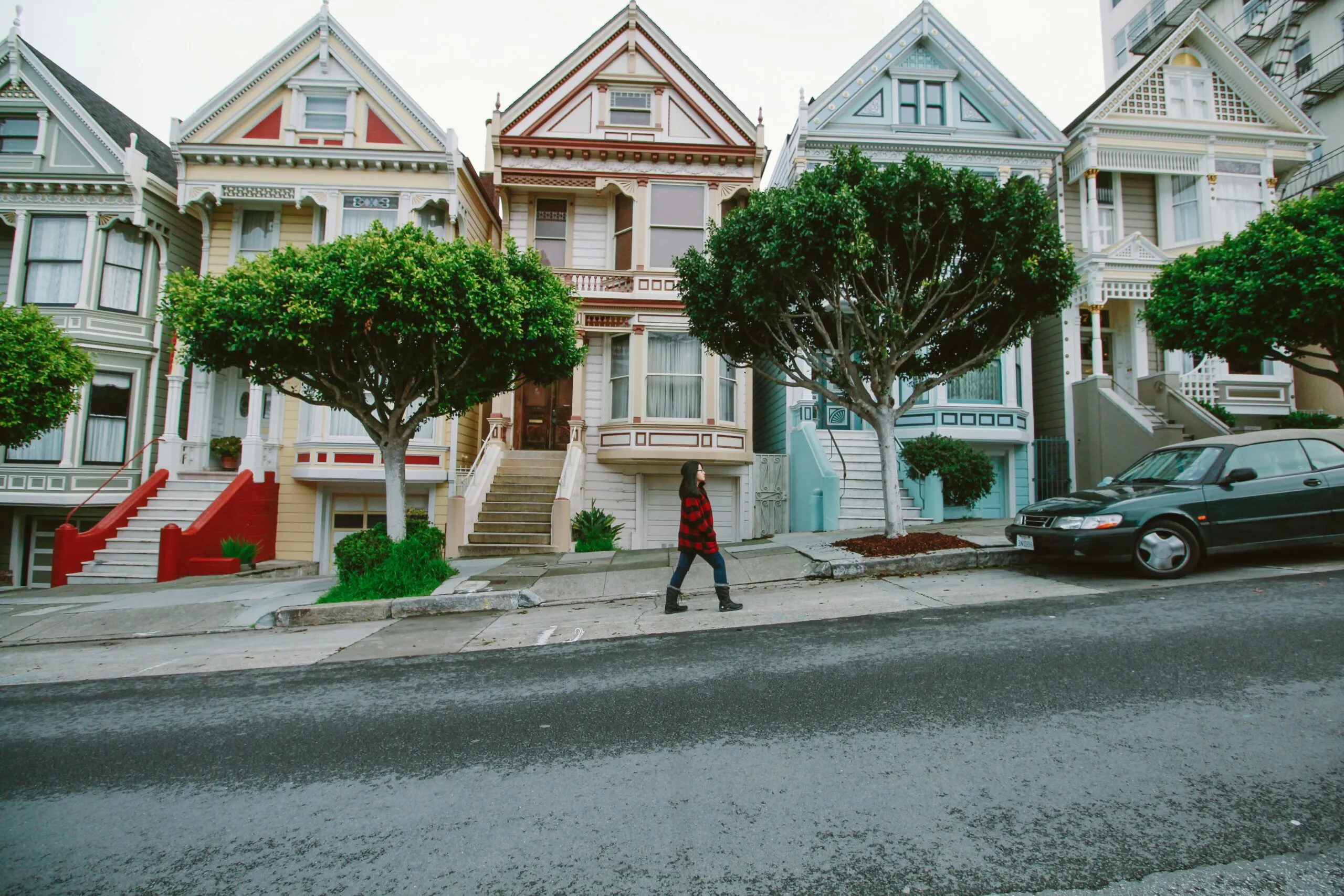 Victorian-style houses line a sloped street, with a person walking and a parked car in view.