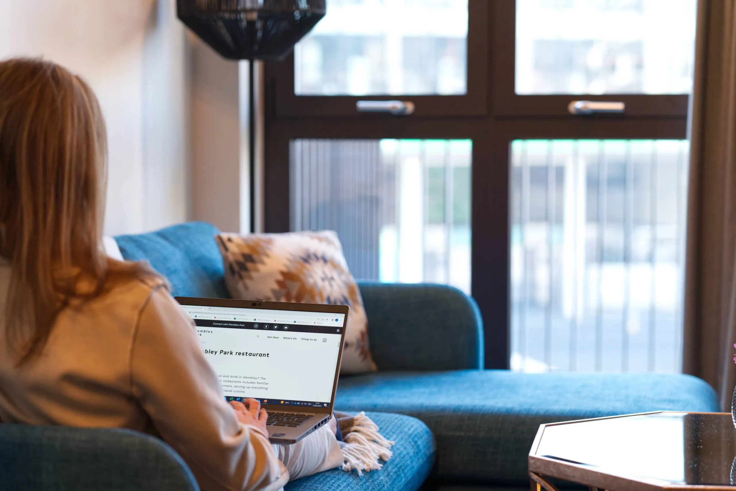 A woman works on a laptop while seated on a blue sofa near a large window in a modern setting.