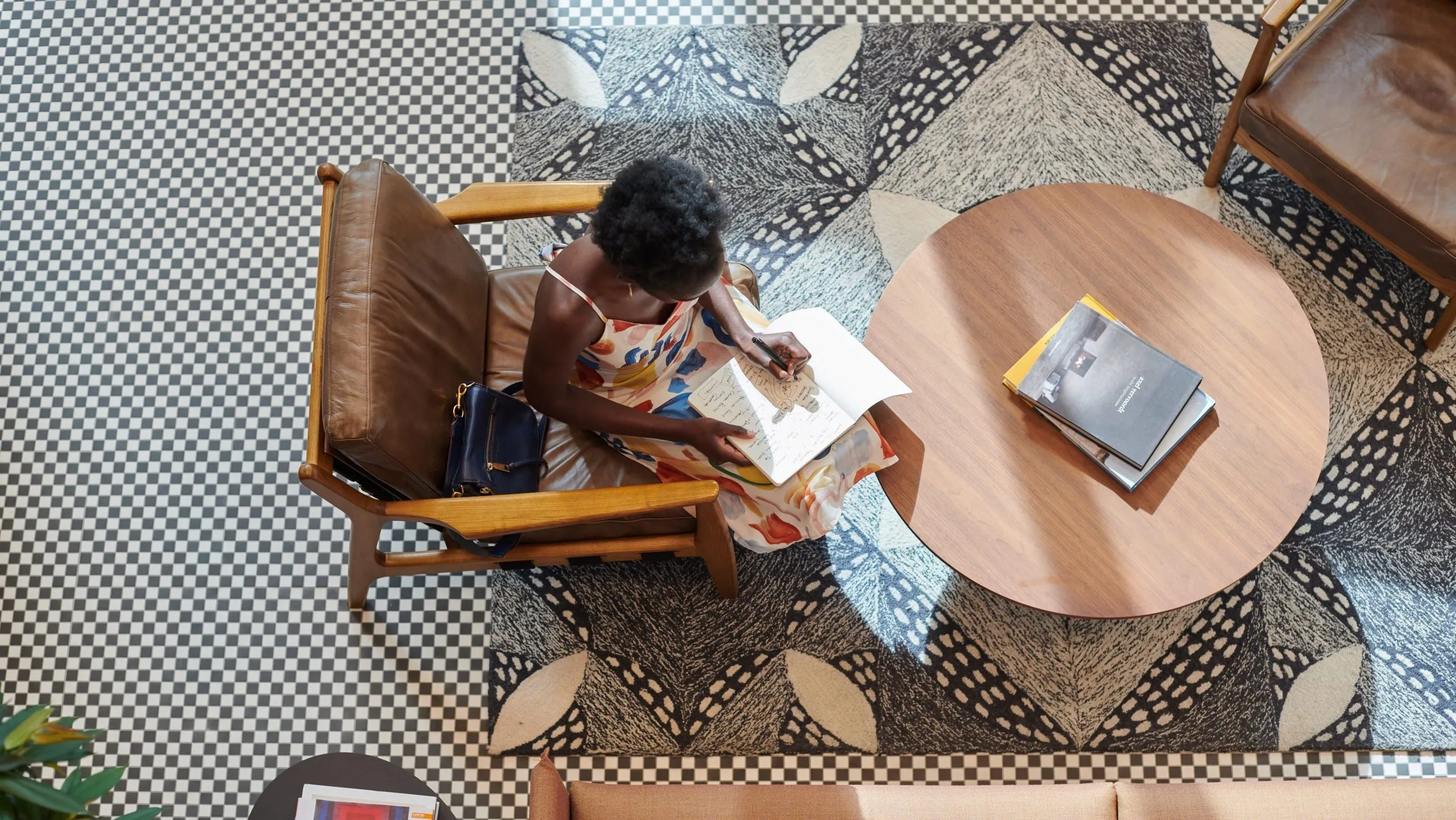 A woman in a floral dress writes in a notebook, seated in a stylish room with patterned flooring.