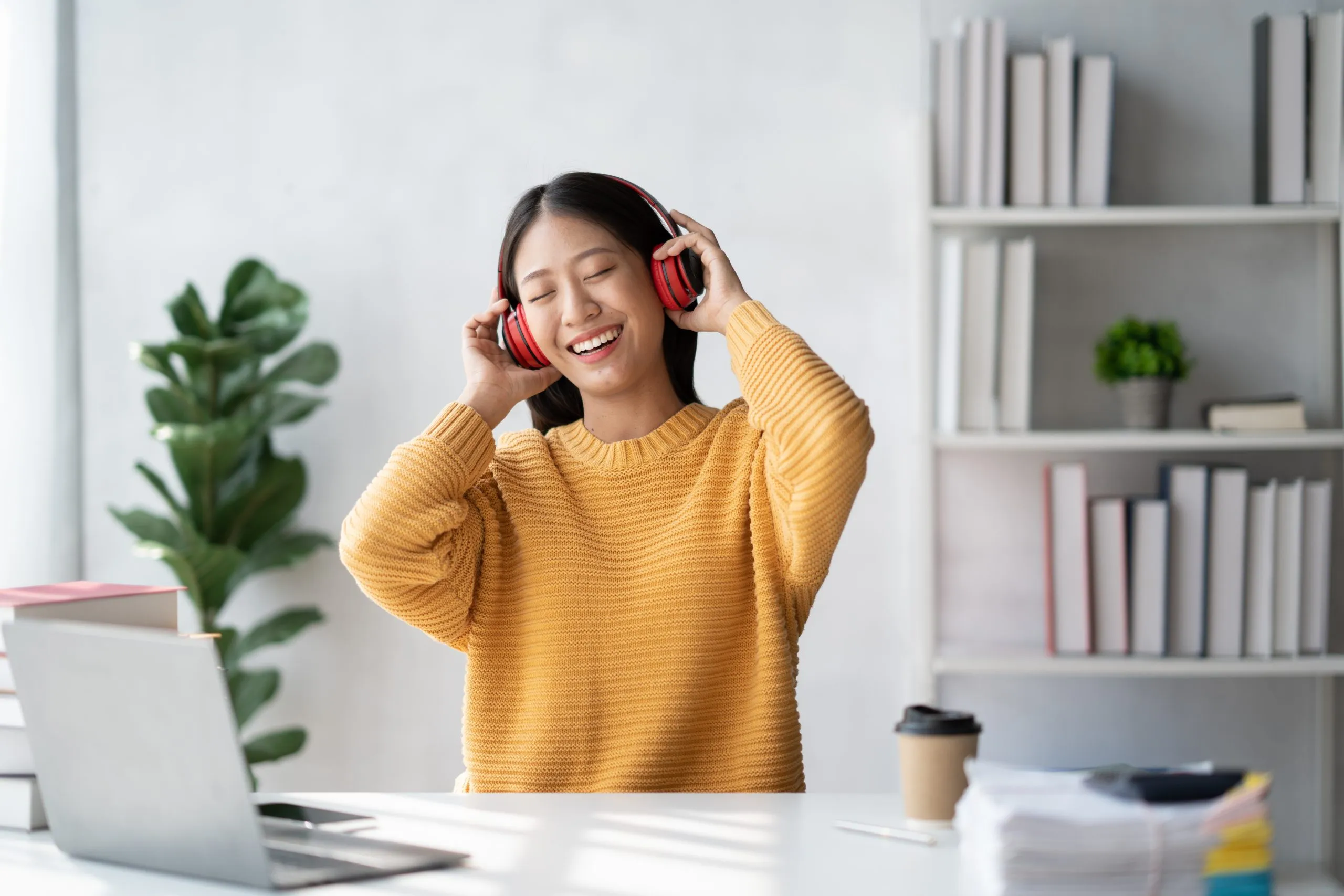 A person wearing a yellow sweater adjusts red headphones while seated at a desk in a bright office.