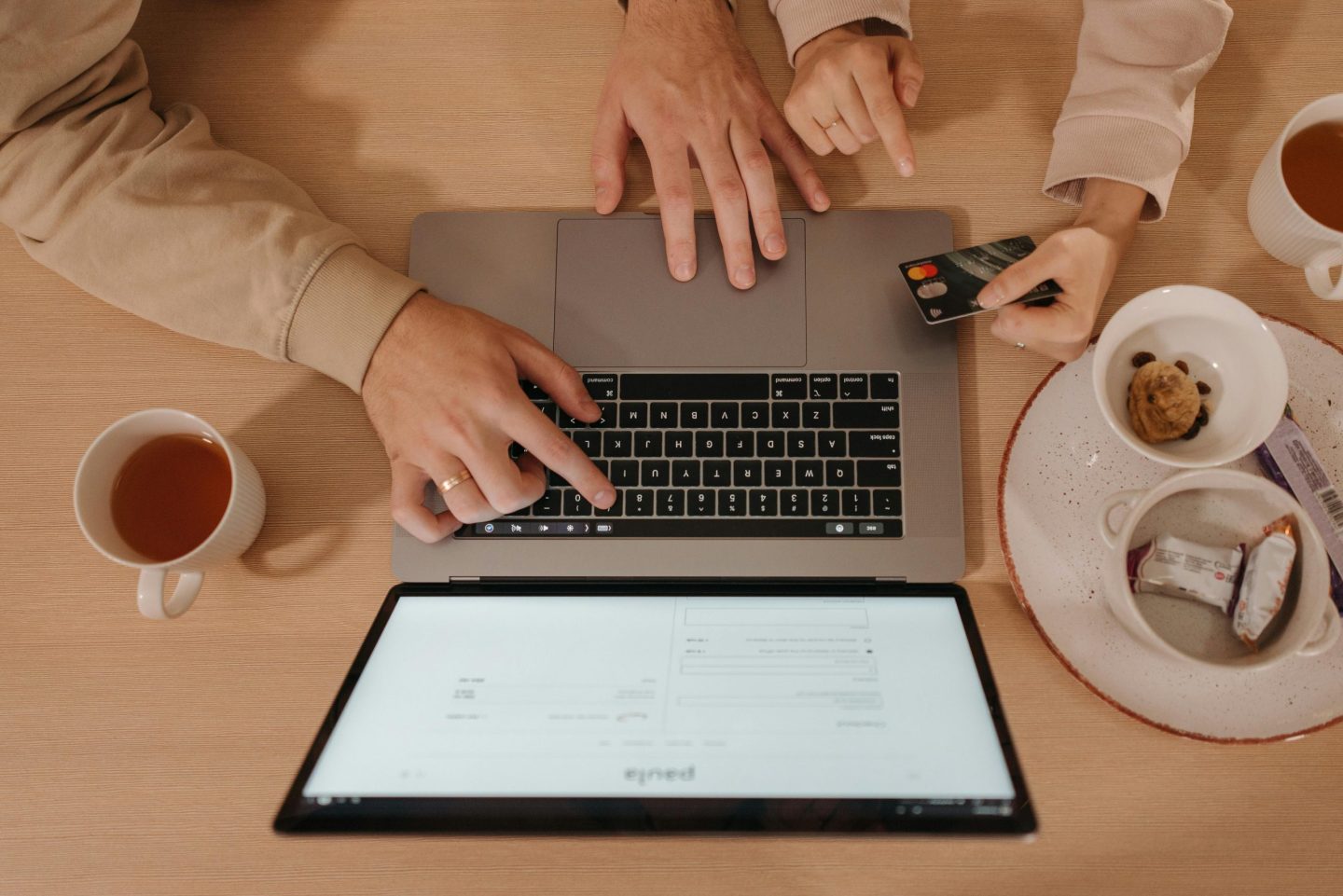 Two people shop online using a laptop, with tea and snacks on a wooden table.