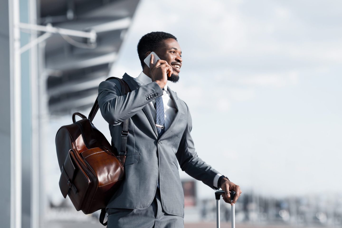 A businessman in a suit with a leather backpack and suitcase talks on the phone outdoors.