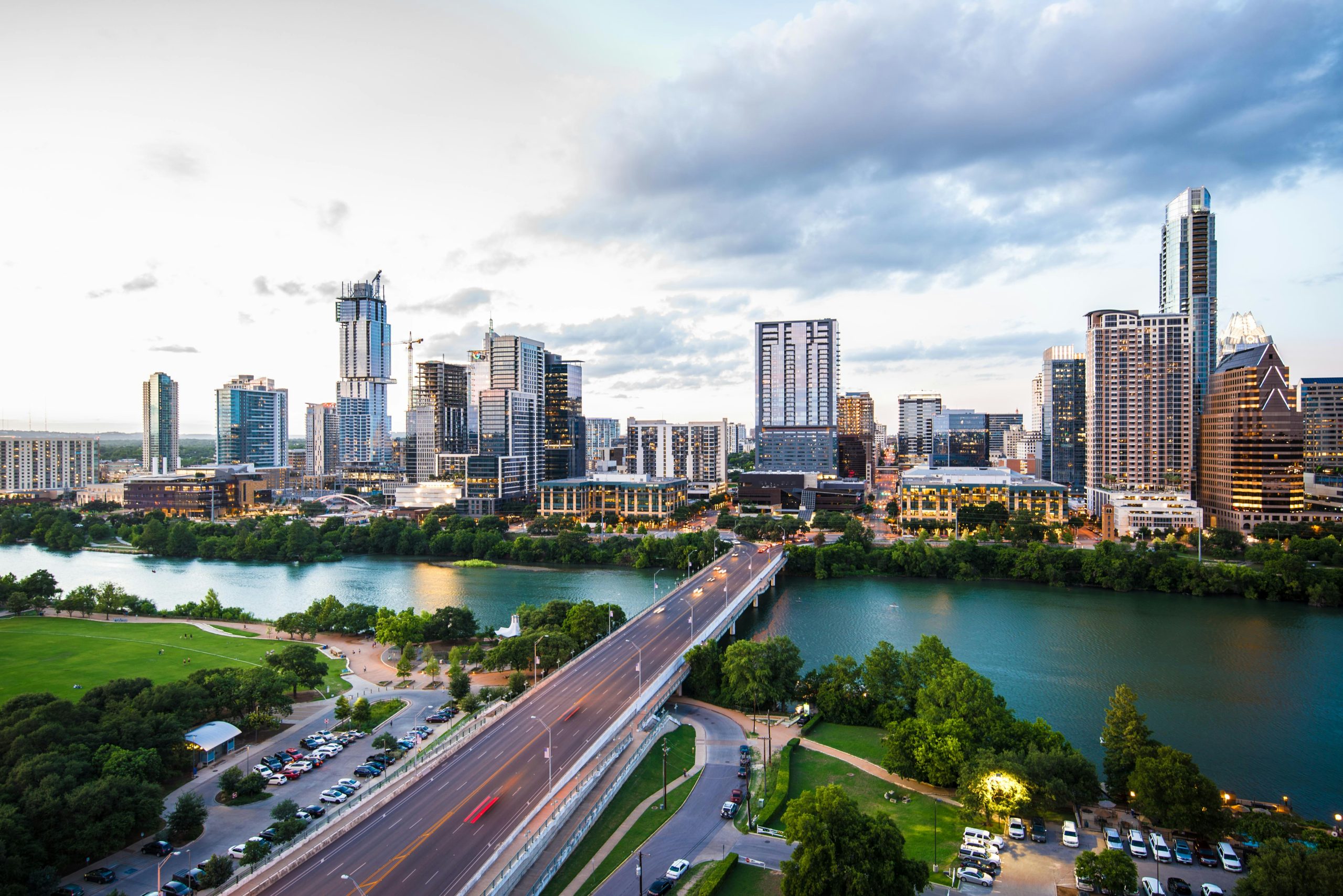 Austin skyline with modern buildings, a river, and a bridge surrounded by greenery at sunset.