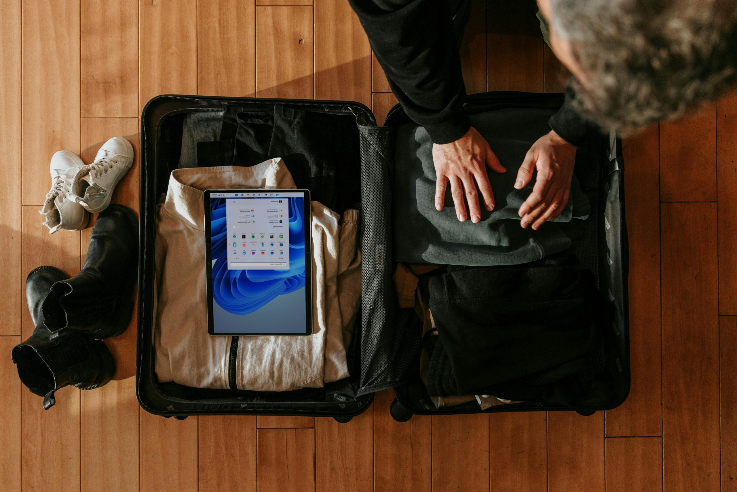 A person packs a suitcase with clothing, a tablet, and shoes on a wooden floor.