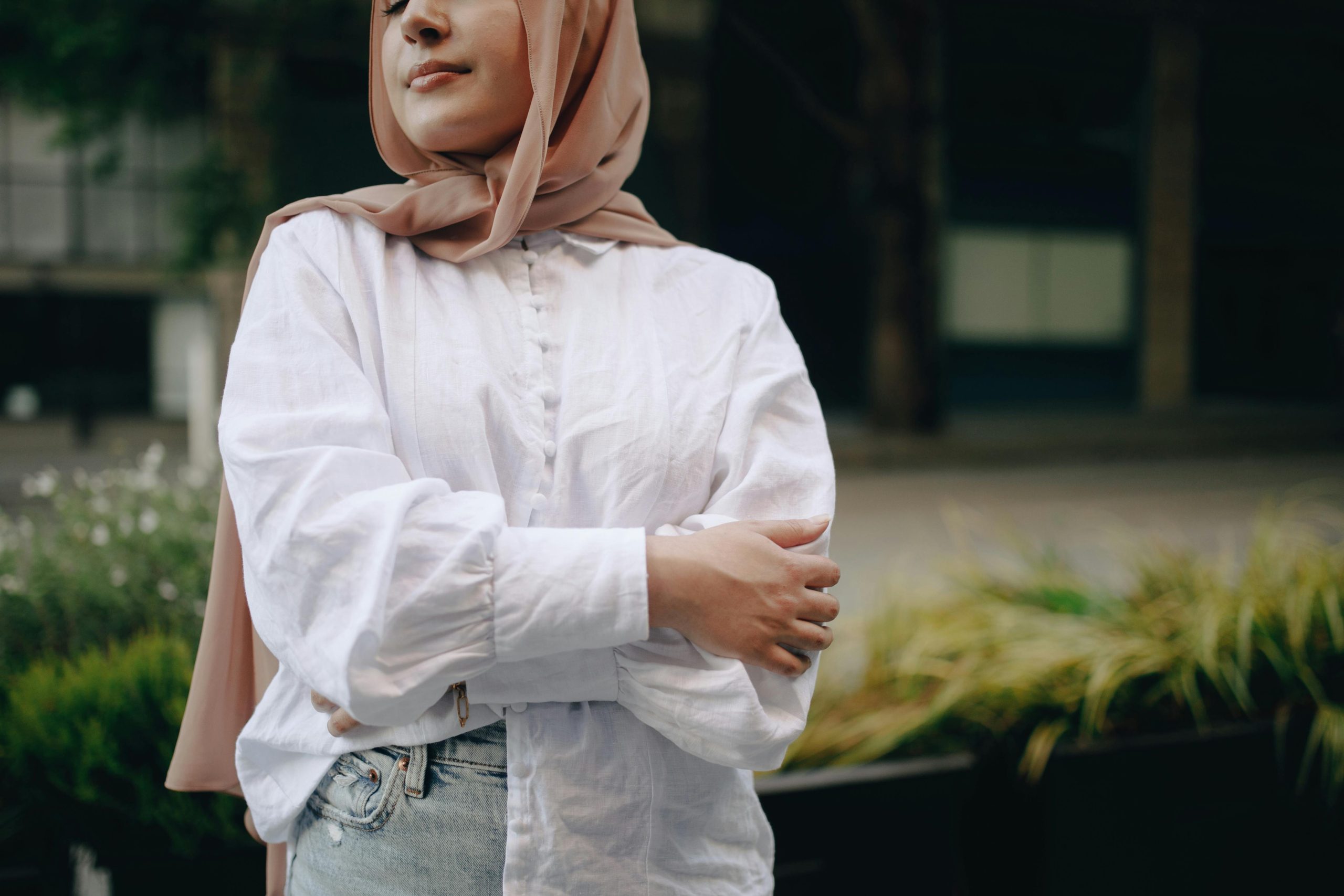 A person wearing a white button-up shirt, light blue jeans, and a beige hijab stands outdoors.