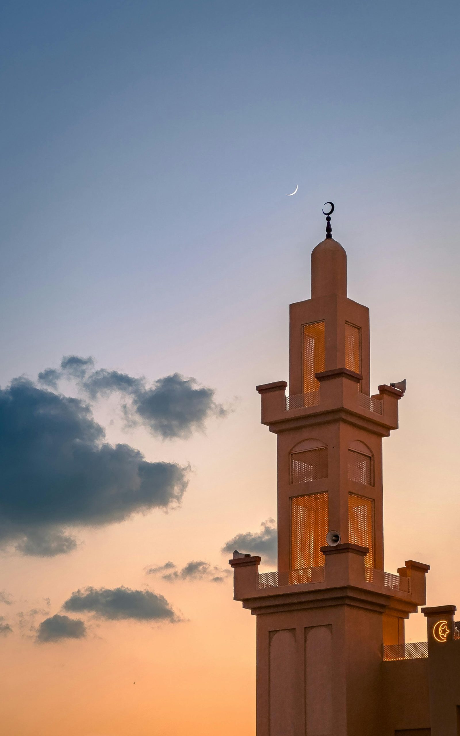 A mosque minaret at sunset with a crescent moon visible in the serene sky.