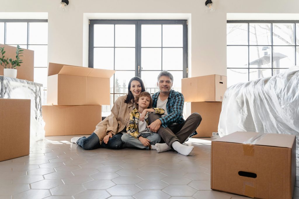 A family sits on the floor of a sunlit room surrounded by moving boxes and wrapped furniture.
