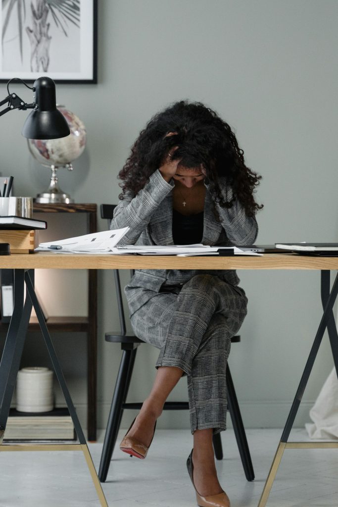 A woman in business attire sits at a desk, holding her head in apparent stress or frustration.