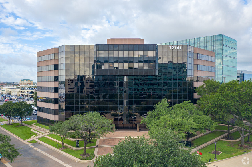 A modern glass office building with reflective windows, surrounded by trees and a paved walkway.