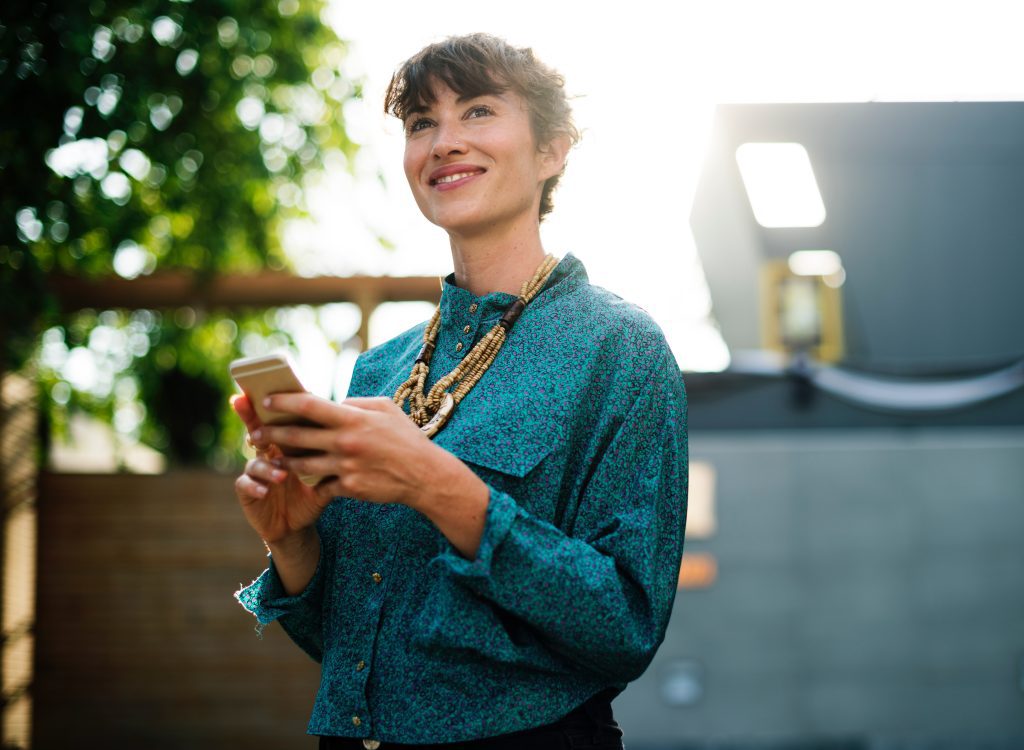 A person wearing a teal patterned shirt and layered necklaces holds a smartphone outdoors.