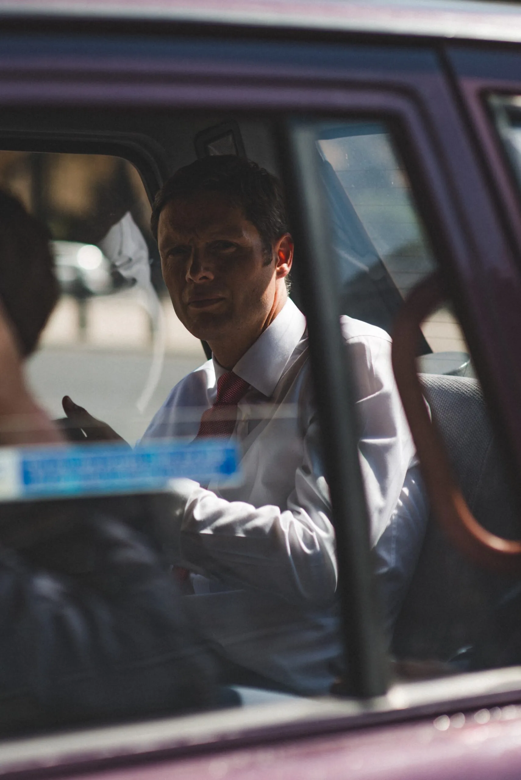 A man in formal attire sits inside a car, partially visible through the window.