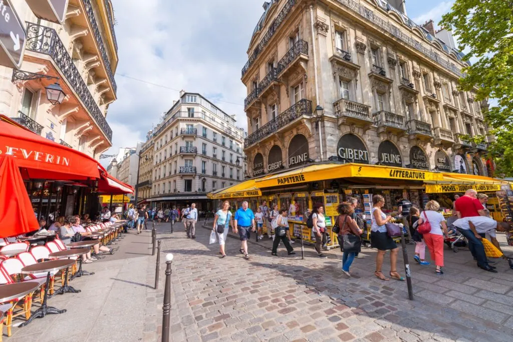 A bustling Parisian street with cafes, bookstores, and pedestrians under a bright, sunny sky.