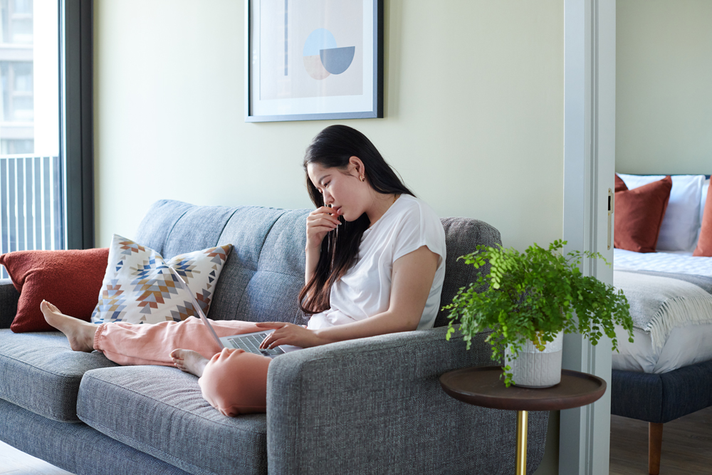 A woman sits on a gray sofa with a laptop, surrounded by modern decor and natural light.