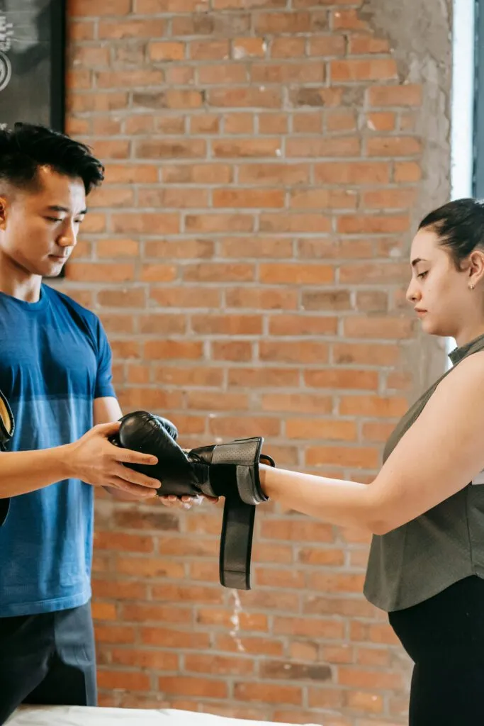 Two individuals exchange boxing gloves in a brick-walled room, suggesting a training or workout session.