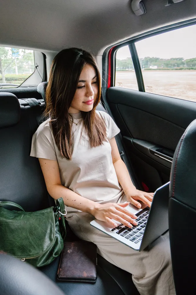 A woman works on a laptop in the backseat of a car, with a green bag and notebook beside her.