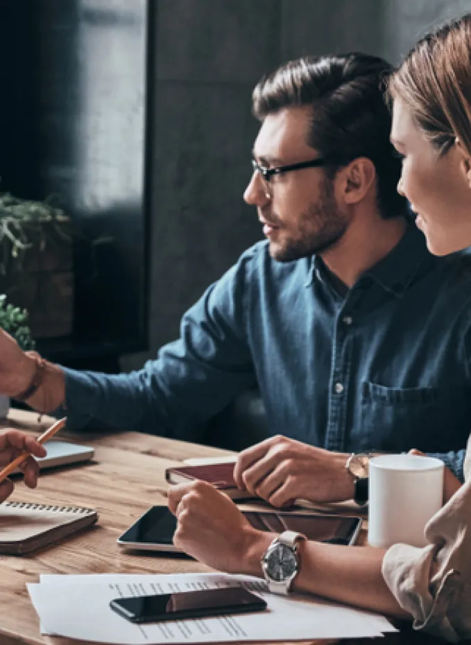 A group collaborates at a wooden table with documents, tablets, notebooks, and coffee mugs.