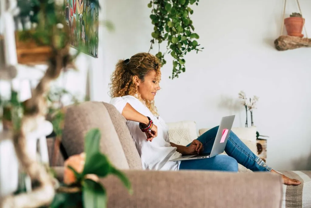 A woman sits on a couch with a laptop, surrounded by plants and a cozy, well-decorated interior.