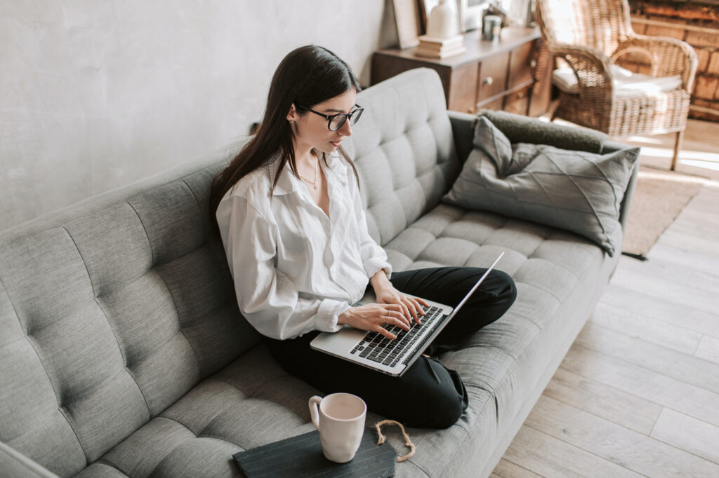 A woman in a white shirt works on a laptop while seated on a gray sofa in a cozy living room.