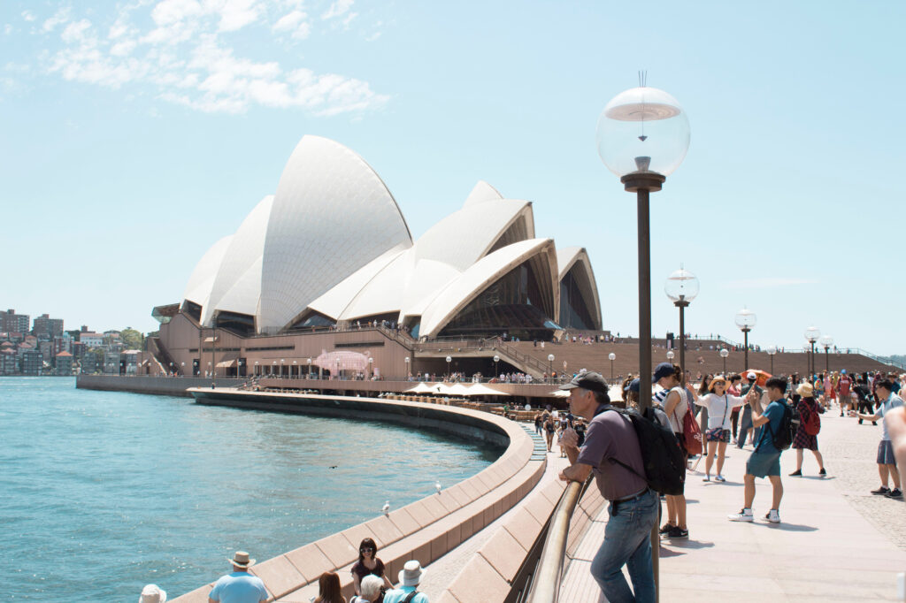 Sydney Opera House with tourists along the waterfront under a clear blue sky.