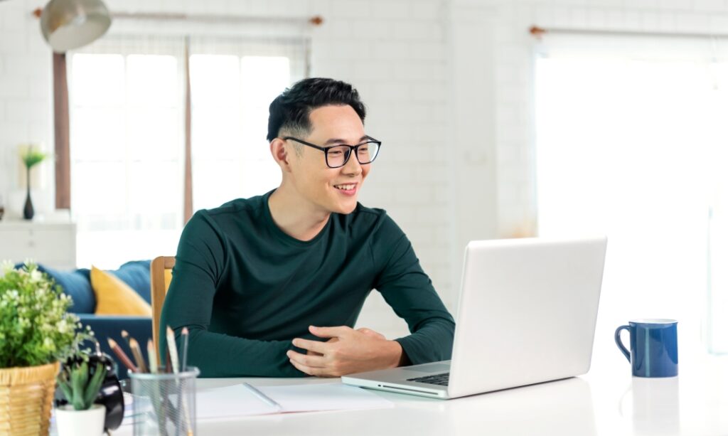 A man wearing glasses sits at a desk with a laptop, surrounded by office supplies and decor.