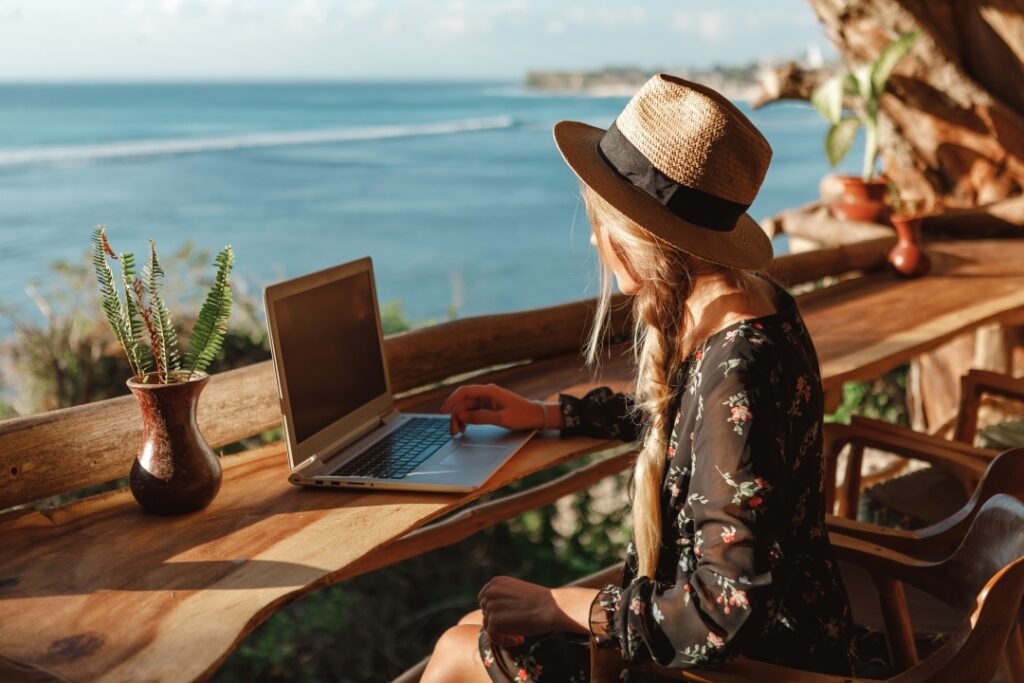 A woman in a straw hat works on a laptop at a wooden desk overlooking the ocean.