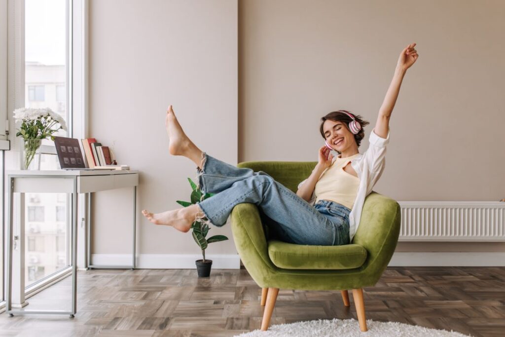 A person relaxes on a green chair, wearing headphones, with legs raised and arm stretched upward.