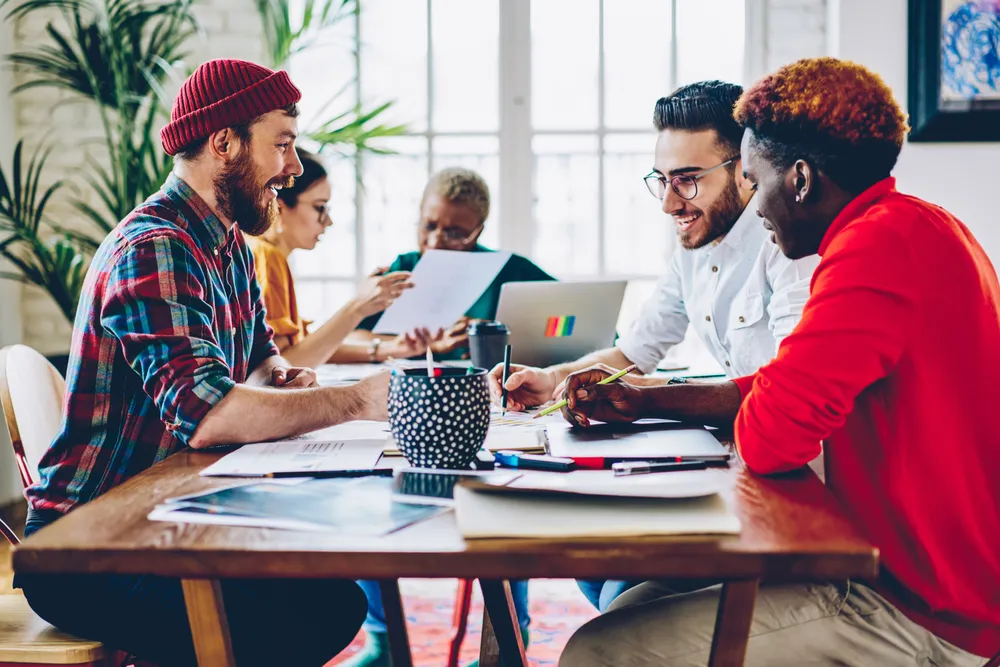 A diverse group collaborates at a table with documents, laptops, and creative materials.