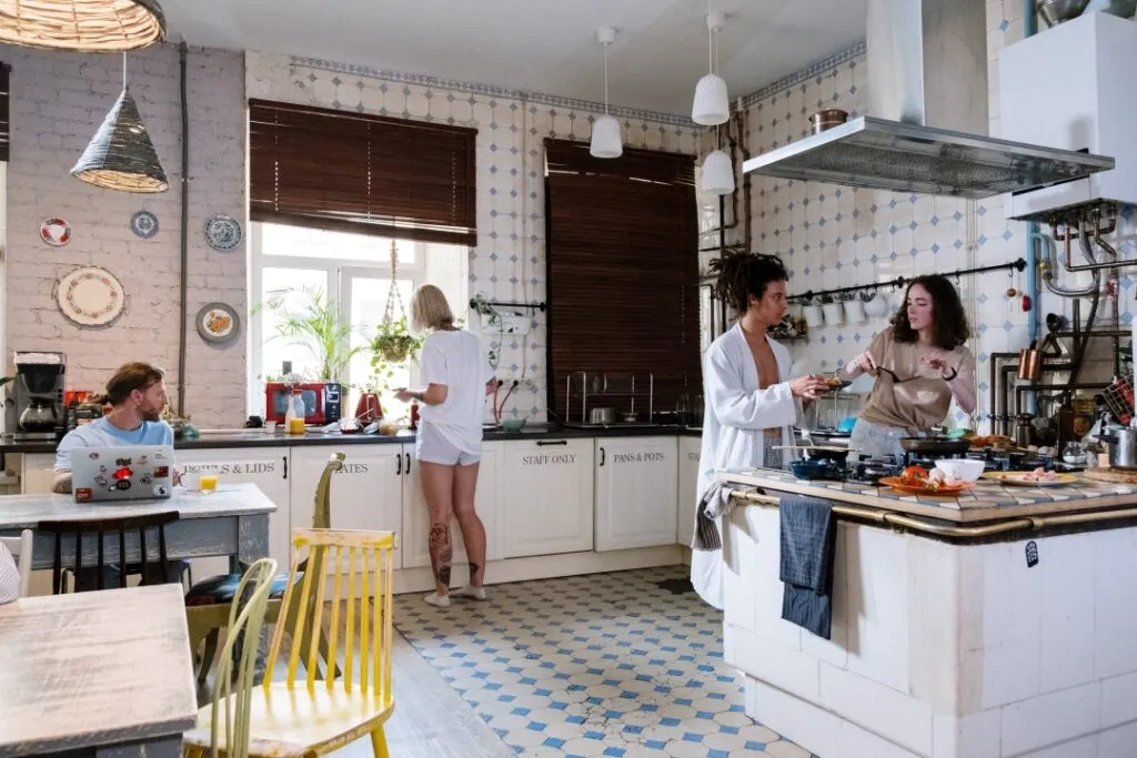A cozy kitchen with people cooking, working, and interacting in a bright, vintage-style setting.