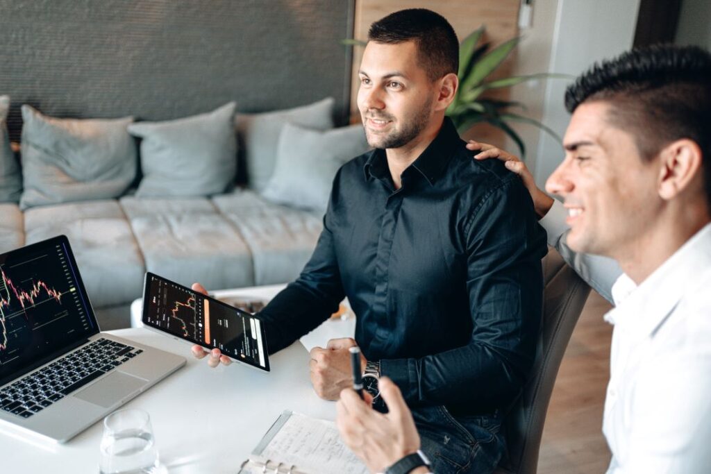 Two individuals discuss stock market data displayed on a laptop and tablet in a modern setting.