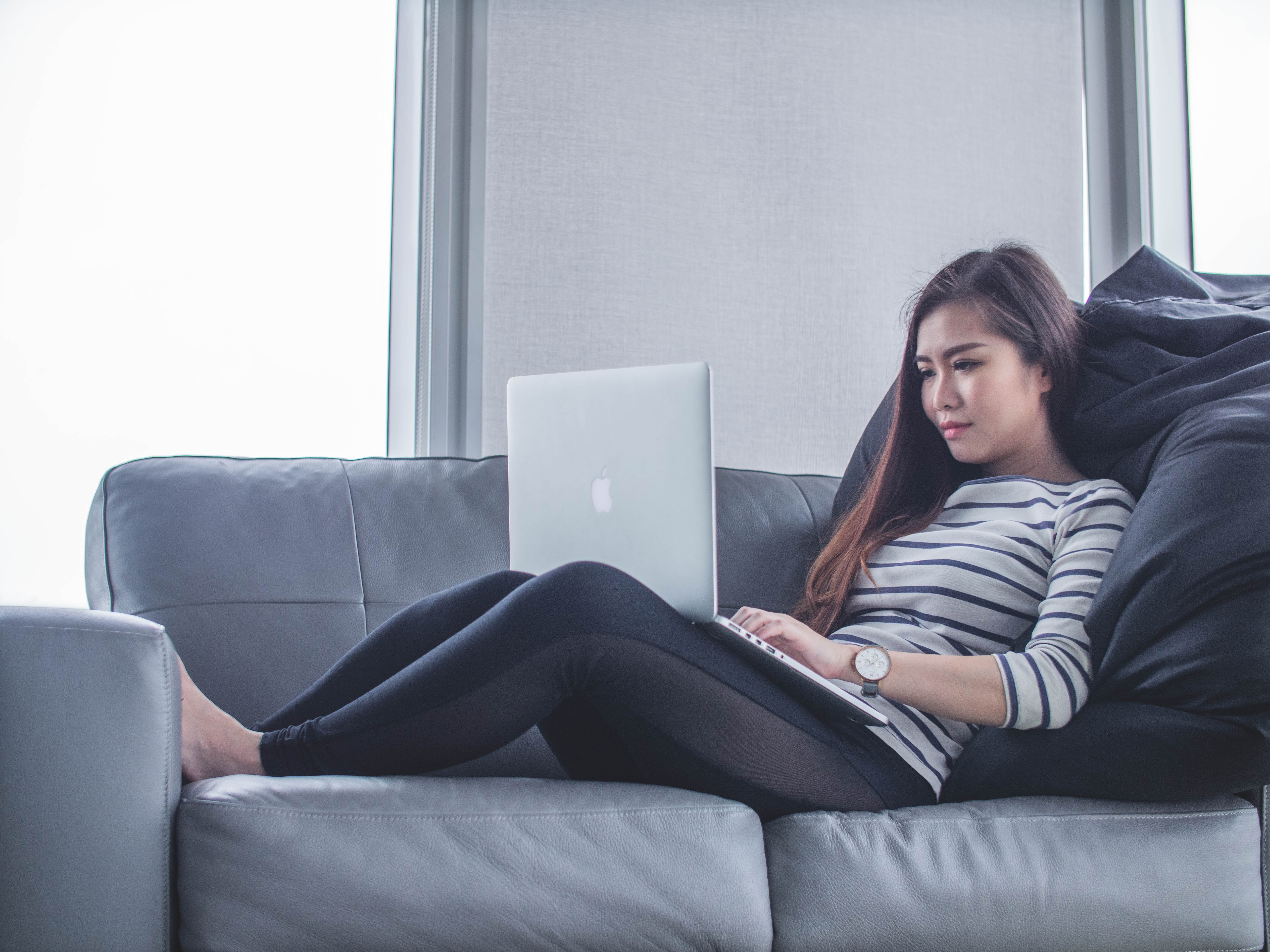 A woman reclines on a sofa, working on a laptop, dressed in a striped top and black leggings.
